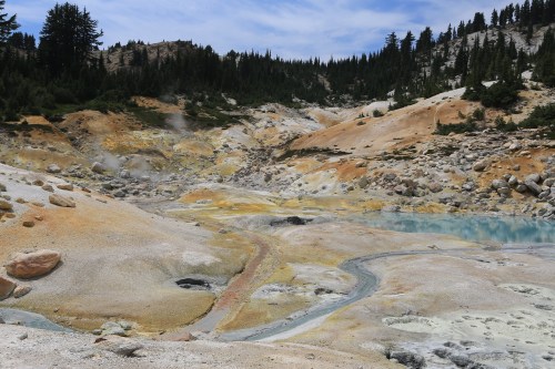A colorful geothermal landscape with steaming pools and rocky terrain, surrounded by trees and mountains under a blue sky.