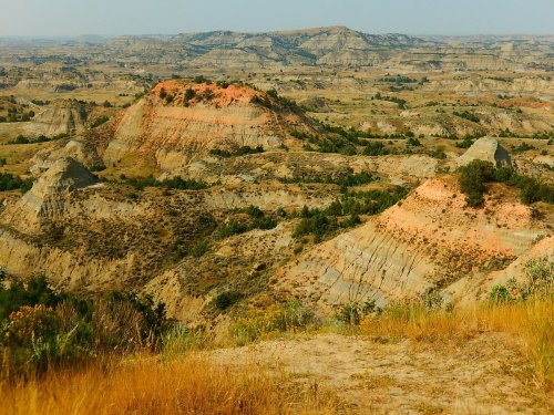 A panoramic view of rugged, colorful hills and valleys under a clear sky, showcasing natural geological formations.