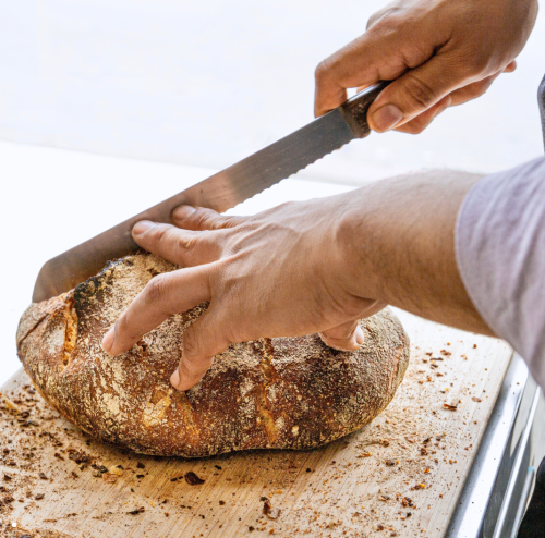 A person slices a round loaf of bread on a wooden cutting board, with crumbs scattered around.