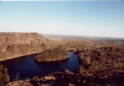 A scenic view of a river with an island, surrounded by trees and hills under a clear blue sky.