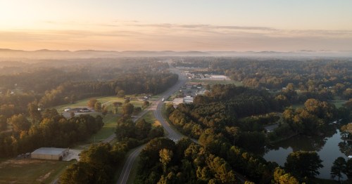Aerial view of a small town surrounded by trees and hills, with a winding road and misty morning light.