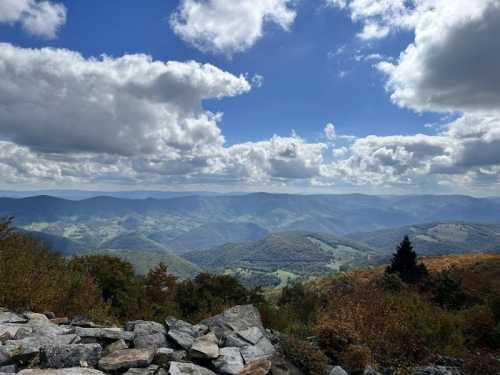 A panoramic view of rolling mountains under a partly cloudy sky, with vibrant autumn foliage in the foreground.