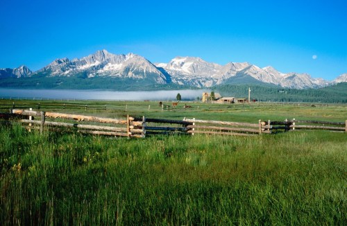 A serene landscape featuring green fields, wooden fences, and majestic mountains under a clear blue sky.