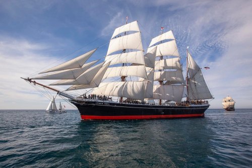 A tall ship with white sails sails on calm waters, with another ship in the background under a blue sky.