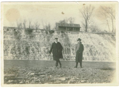 Two men stand by a waterfall, with a wooden structure visible in the background, in a vintage black-and-white photo.