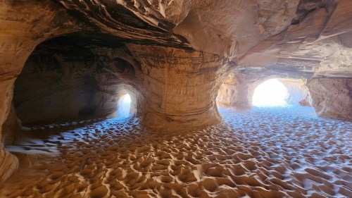 A sandy cave interior with light streaming through openings, highlighting textured rock formations.