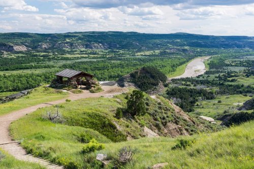 A scenic view of a green valley with a winding river, featuring a small cabin on a hilltop under a cloudy sky.