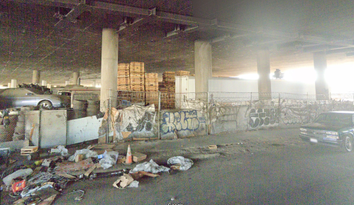 A dimly lit urban area under a bridge, featuring graffiti, discarded items, and stacked wooden pallets.