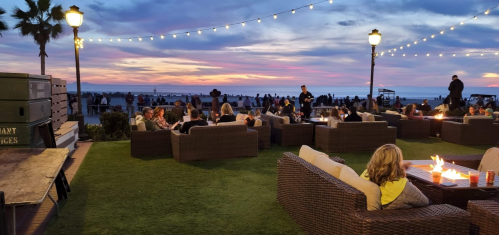 A lively outdoor gathering at sunset, with people seated around fire pits and string lights illuminating the scene.