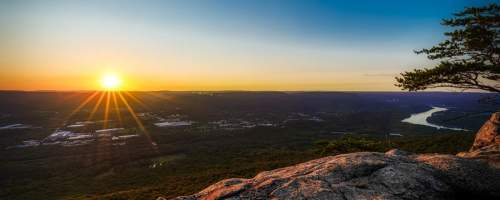 Sunset over a valley, with a river winding through lush greenery and rocky foreground, creating a serene landscape.