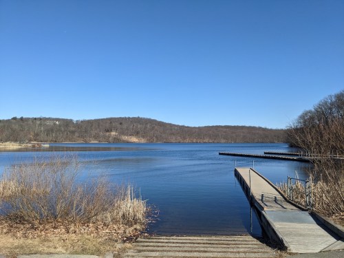 A serene lake scene with a wooden dock, surrounded by trees and hills under a clear blue sky.