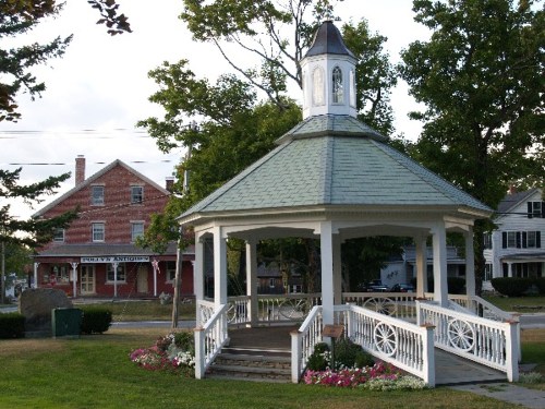 A white gazebo with a green roof surrounded by trees and flowers, with a historic building in the background.