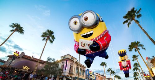 A large Minion balloon smiles in a festive setting, with palm trees and a nutcracker balloon in the background.