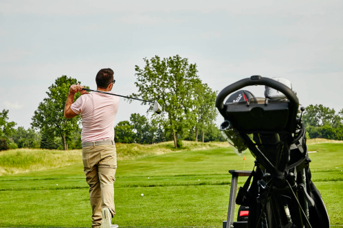 A golfer swings a club on a lush green course, with a golf bag in the foreground.