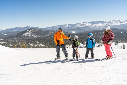 A family of four stands on a snowy slope, ready to ski, with mountains in the background and clear blue skies.
