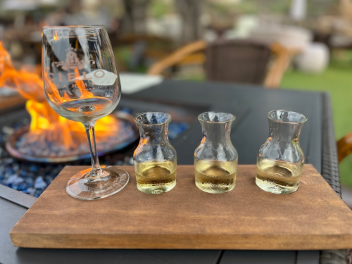 A wine glass beside three small glass carafes of yellow liquid on a wooden board, with a fire pit in the background.