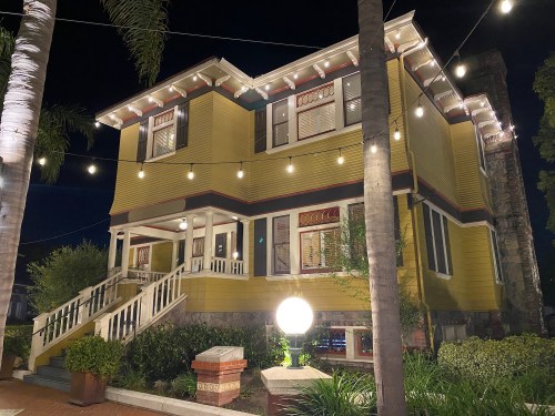 A yellow two-story house at night, illuminated by string lights, surrounded by palm trees and greenery.