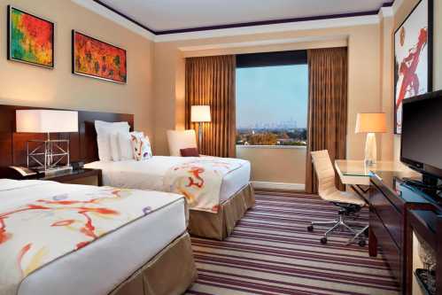 A stylish hotel room with two beds, modern decor, a desk, and a large window showcasing a city view.