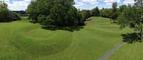 A panoramic view of lush green hills with winding paths and trees under a bright blue sky with fluffy clouds.