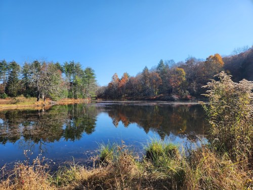 A serene lake surrounded by autumn foliage, reflecting trees and a clear blue sky.