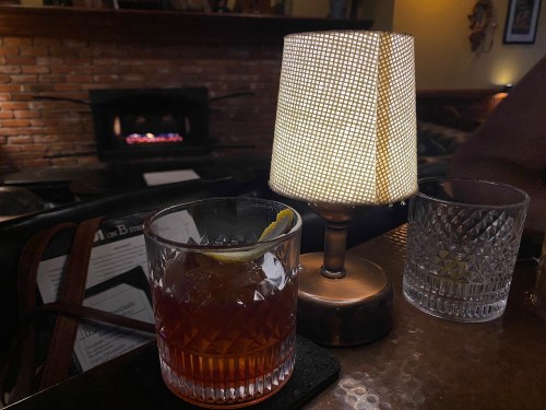 A dimly lit table with a cocktail glass, a lamp, and a cozy fireplace in the background.