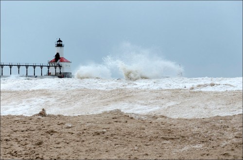 A lighthouse stands on a pier as large waves crash against the shore under a cloudy sky.
