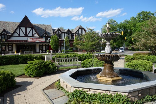 A decorative fountain in a park surrounded by greenery and benches, with a charming building in the background.