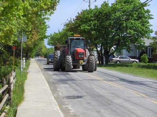 A red tractor drives down a rural road lined with trees and houses, with a car following behind.