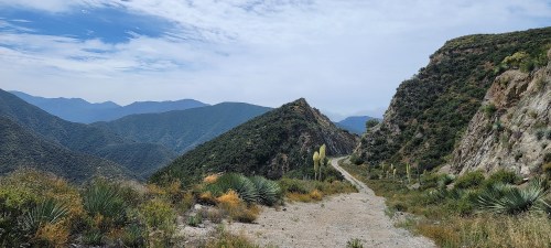 A winding dirt path through lush green mountains under a cloudy sky.