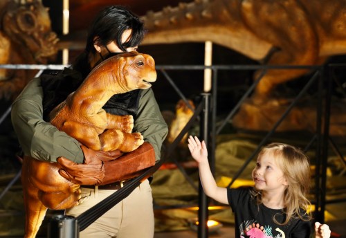 A child smiles and waves at an adult holding a dinosaur puppet in a museum exhibit.