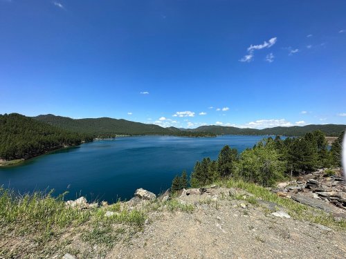 A serene lake surrounded by green hills and trees under a clear blue sky with scattered clouds.