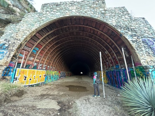 A person stands at the entrance of a large, graffiti-covered tunnel, with a bright light visible at the far end.