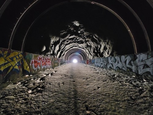 A dark tunnel with graffiti on the walls, leading to a bright light at the far end. The ground is uneven and dusty.