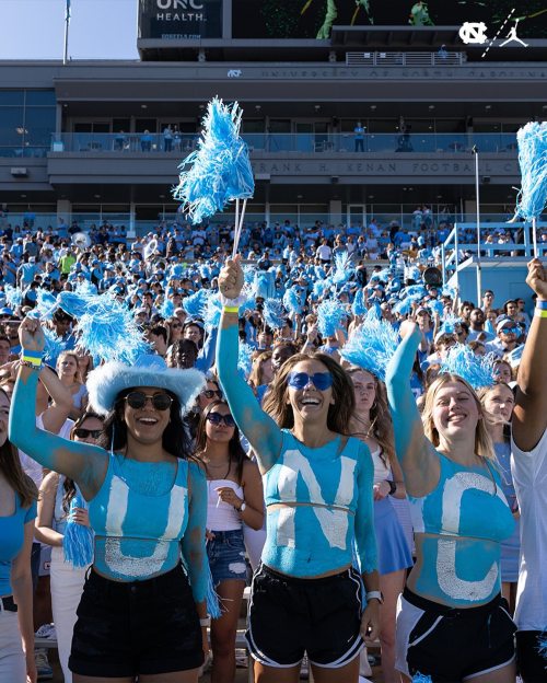 Three fans in blue body paint cheer with pom-poms at a UNC football game, surrounded by a lively crowd.