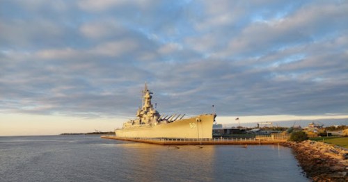 A battleship docked by the shore under a cloudy sky, with calm waters reflecting the scene.