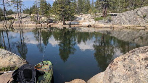 A serene lake surrounded by rocks and trees, with a kayak resting on the shore and reflections in the water.