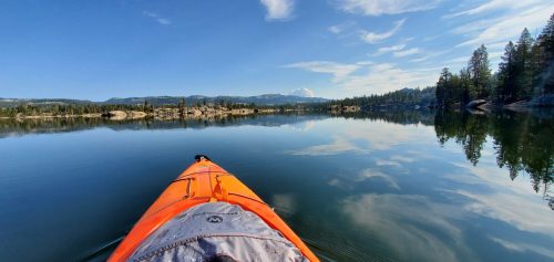 A kayak on calm water, reflecting trees and mountains under a clear blue sky with wispy clouds.