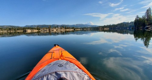 A bright orange kayak on calm water, surrounded by trees and mountains under a clear blue sky.