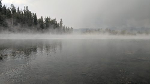 A misty lake surrounded by trees, with fog hovering over the water and a serene, tranquil atmosphere.