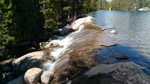 A serene lakeside scene with a gentle waterfall flowing over rocks, surrounded by trees and calm water.