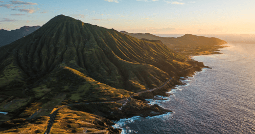 Aerial view of a lush green mountain beside a coastline at sunset, with waves crashing against the shore.