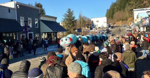A crowd gathers on a street for a parade featuring a large blue fish puppet, with buildings and trees in the background.