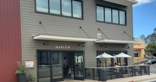 Exterior of a modern building with large windows, featuring the name "Marlow" and outdoor seating with umbrellas.