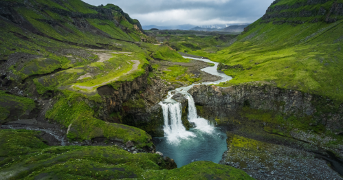 Aerial view of a lush green landscape with a cascading waterfall and winding river surrounded by mountains.