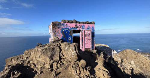 A colorful, graffiti-covered structure on a rocky cliff overlooking the ocean under a clear blue sky.