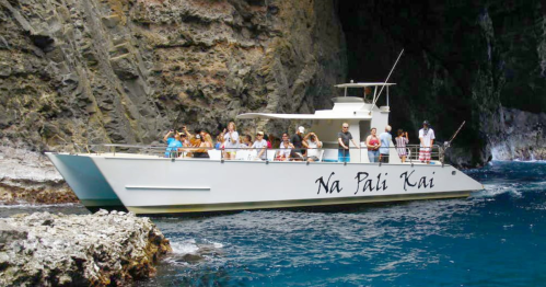 A tour boat named "Na Pali Kai" with passengers cruising along a rocky coastline and clear blue waters.