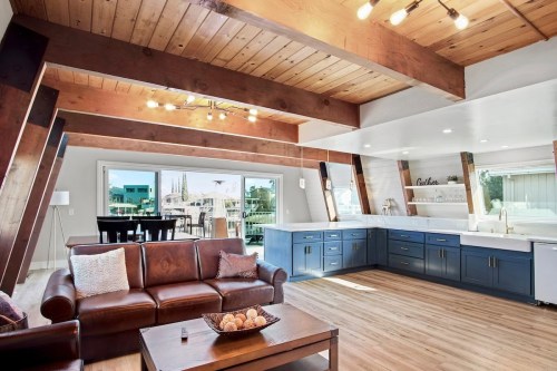 Bright, modern living space with wooden beams, a leather sofa, and a kitchen featuring blue cabinets and large windows.
