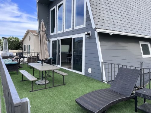 A modern rooftop terrace with a table, chairs, and lounge chair on artificial grass, featuring large windows and a clear sky.