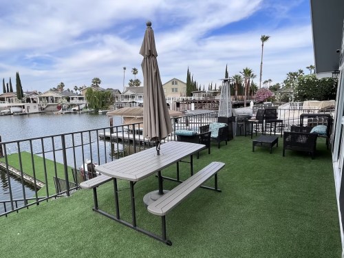 A patio with a table and chairs overlooking a calm lake, surrounded by palm trees and houses.