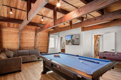 A cozy game room featuring a blue pool table, foosball table, and wooden beams, with a sofa and wall-mounted TV.